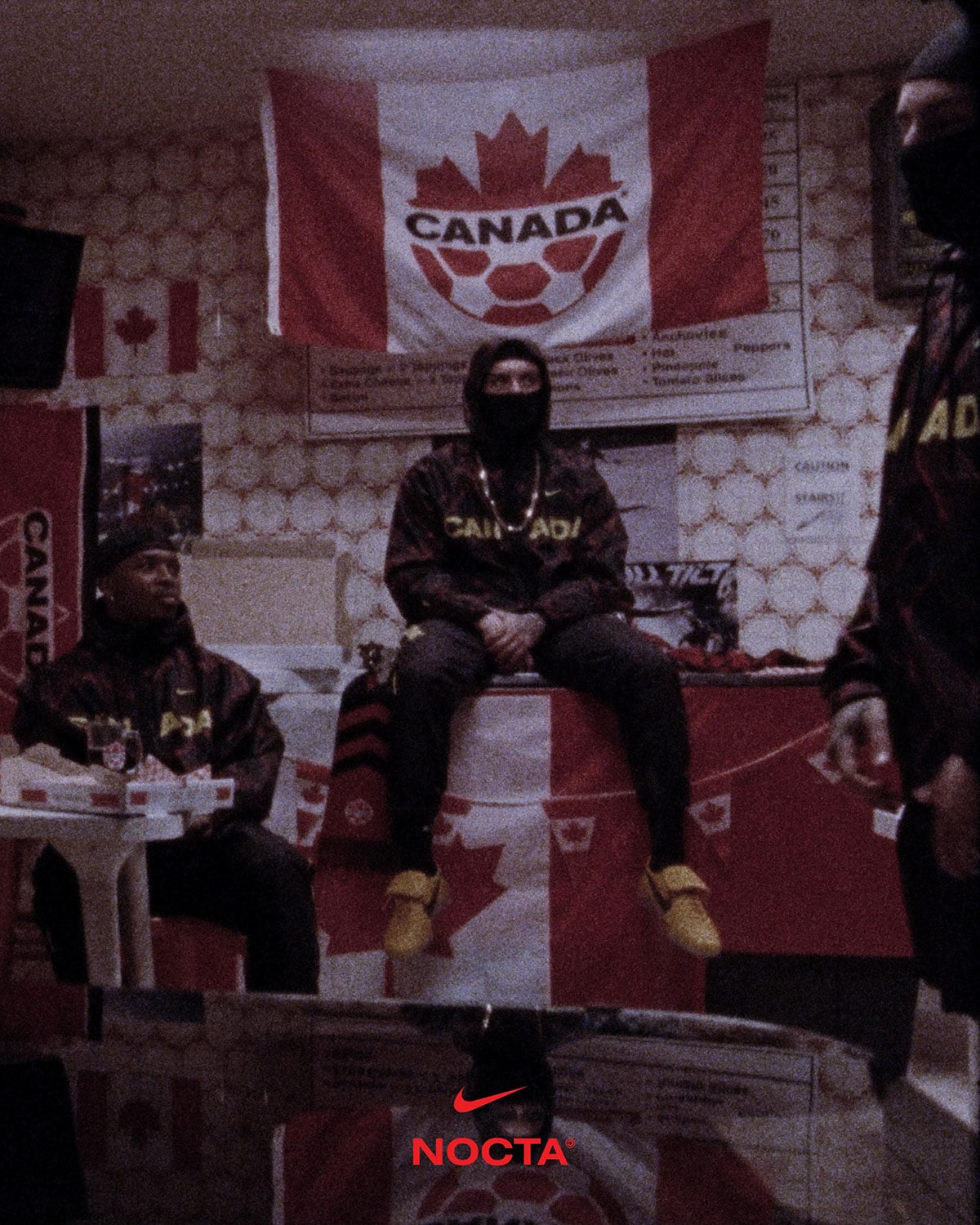 Group seated under a Canada Soccer flag, the center figure wearing the wheat NOCTA Nike Cryoshot on foot