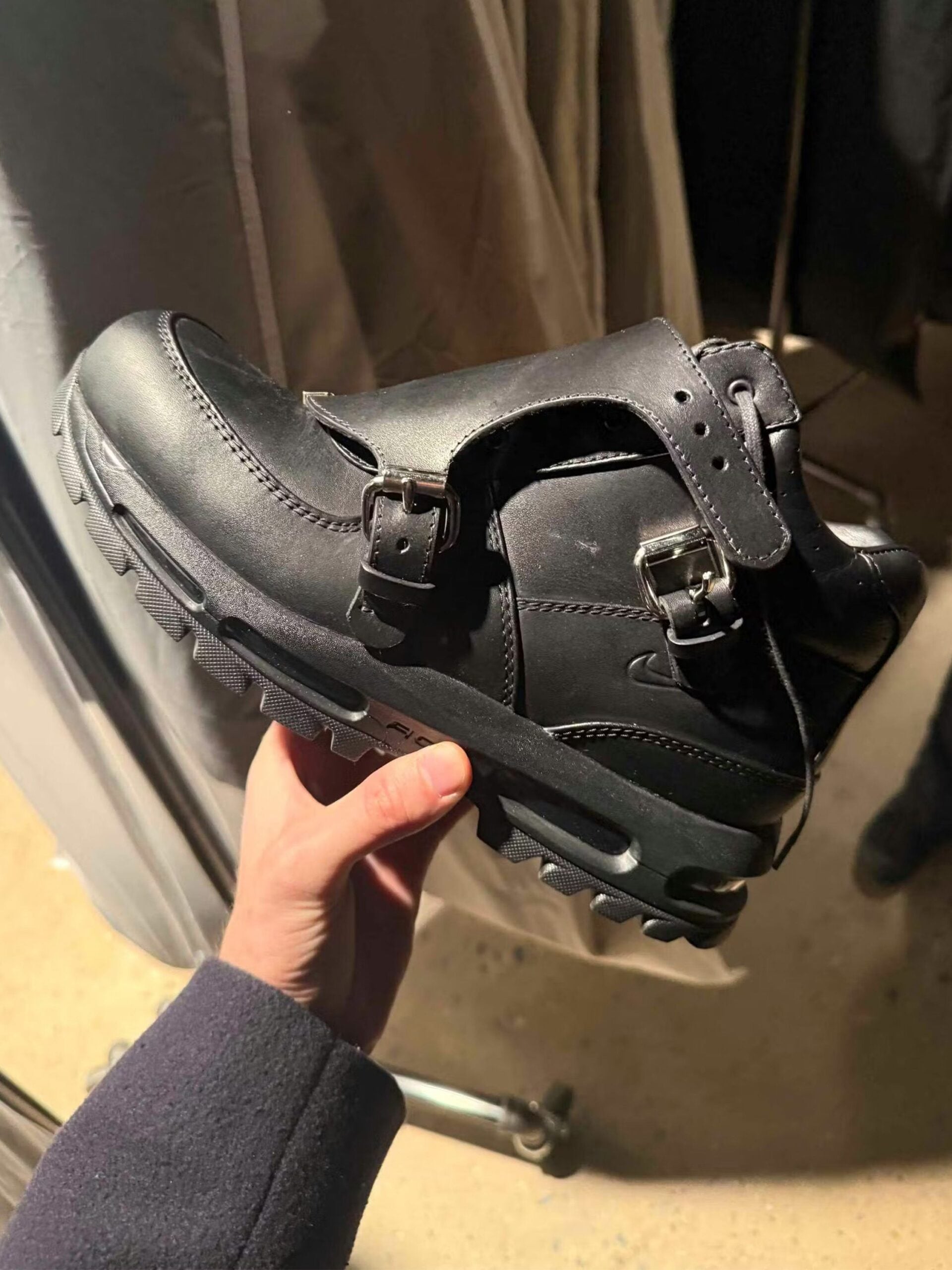Heel and medial view of the PSNY Air Max Goadome held in hand, twin buckle straps over premium black leather