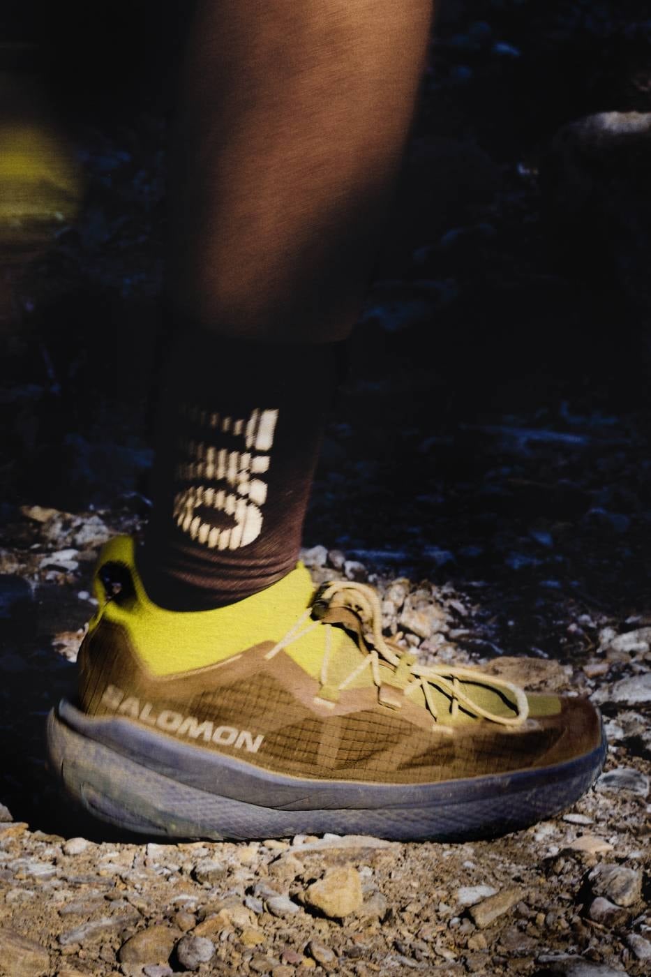 Low-angle look at the olive and yellow GRVL Concept showing the supercritical foam midsole on a rocky riverbank