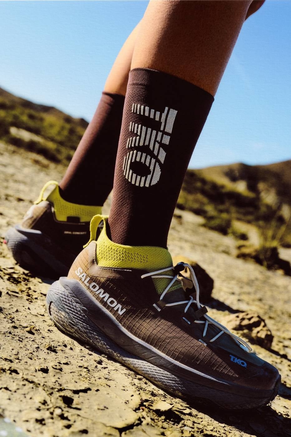 On-foot side profile of the GRVL Concept gravel-running shoe with a yellow knit gaiter and blue accents on rocky terrain