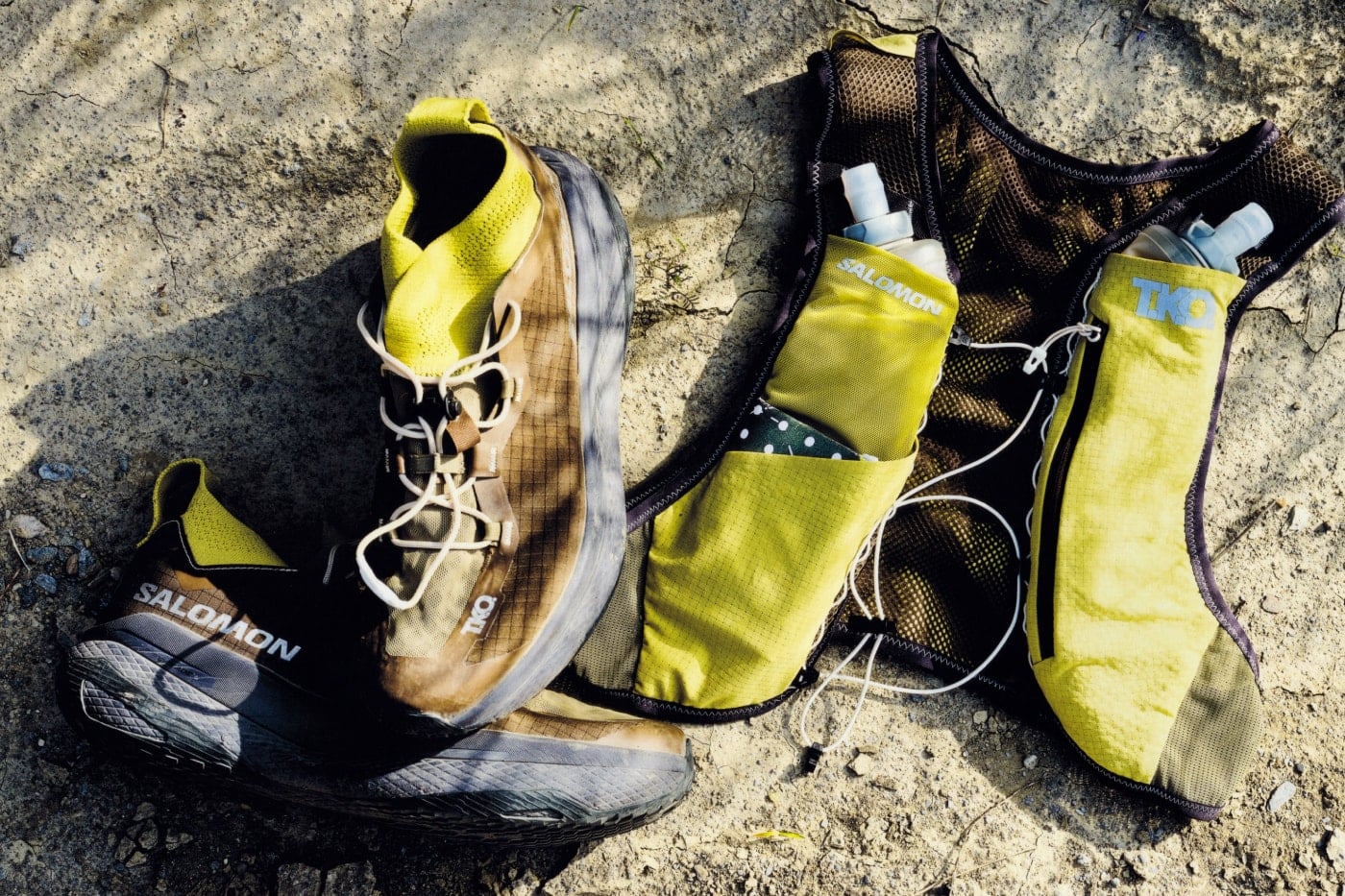Flat-lay of the Salomon Pas Normal Studios GRVL Concept shoe and RACE SKIN 2 vest on cracked, sandy ground