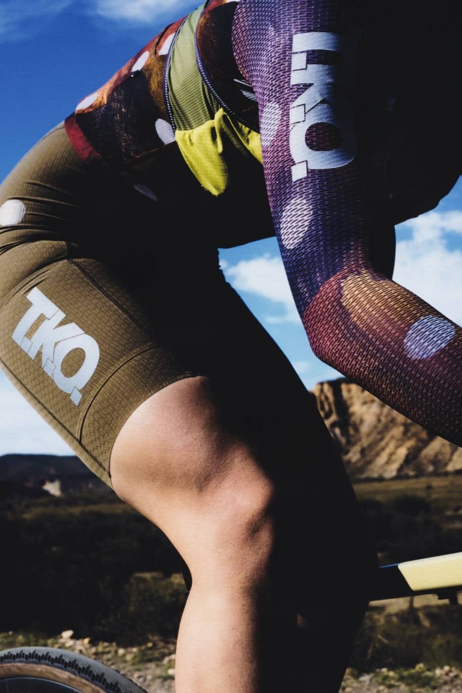 Cyclist riding gravel in the T.K.O. Pas Normal Studios cycling kit under a blue sky, the capsule crossover idea in action