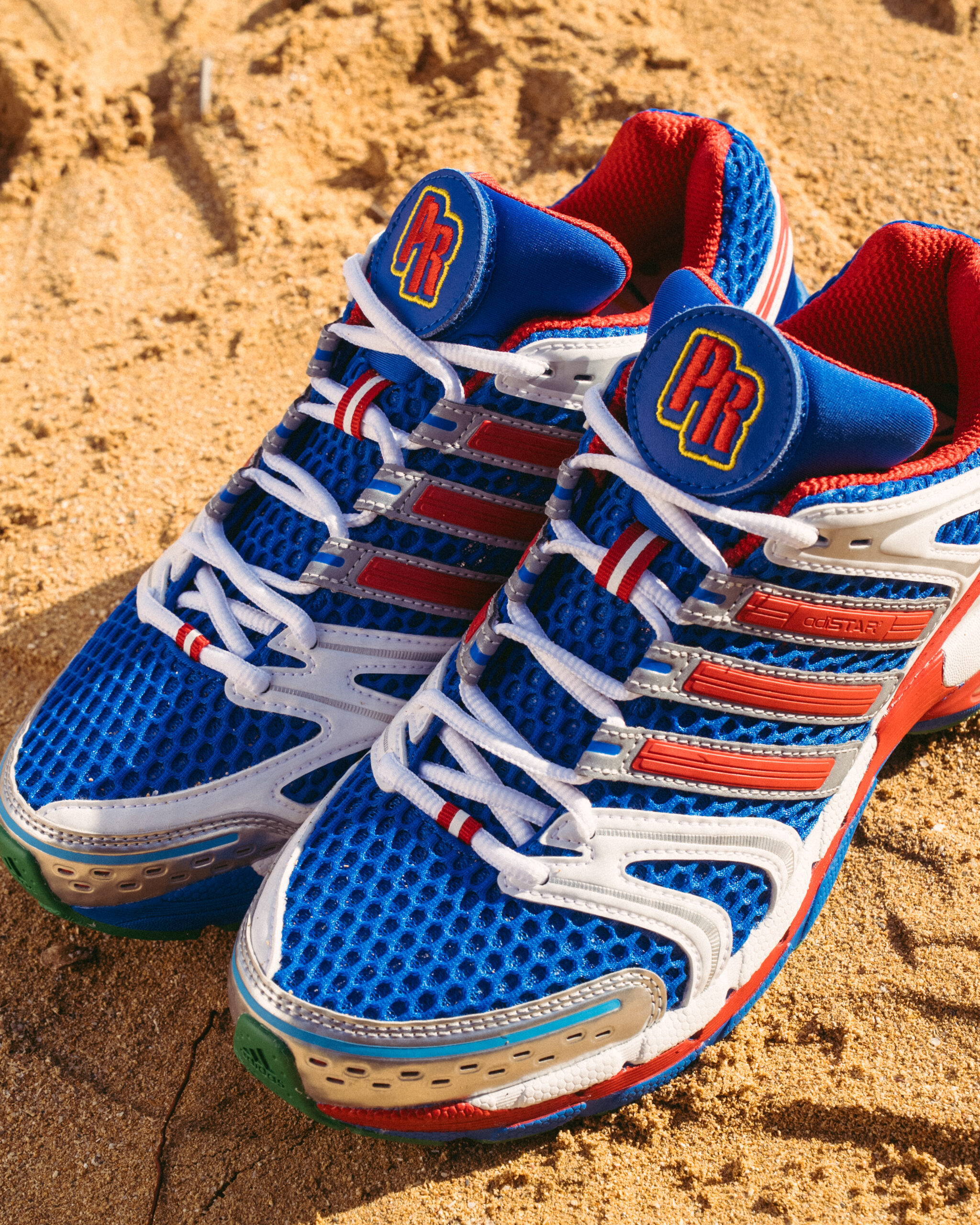 Pair of the Sneaker Room x adidas Adistar Control 5 "PR" on sand, with the PR tongue patch in red, white, and blue