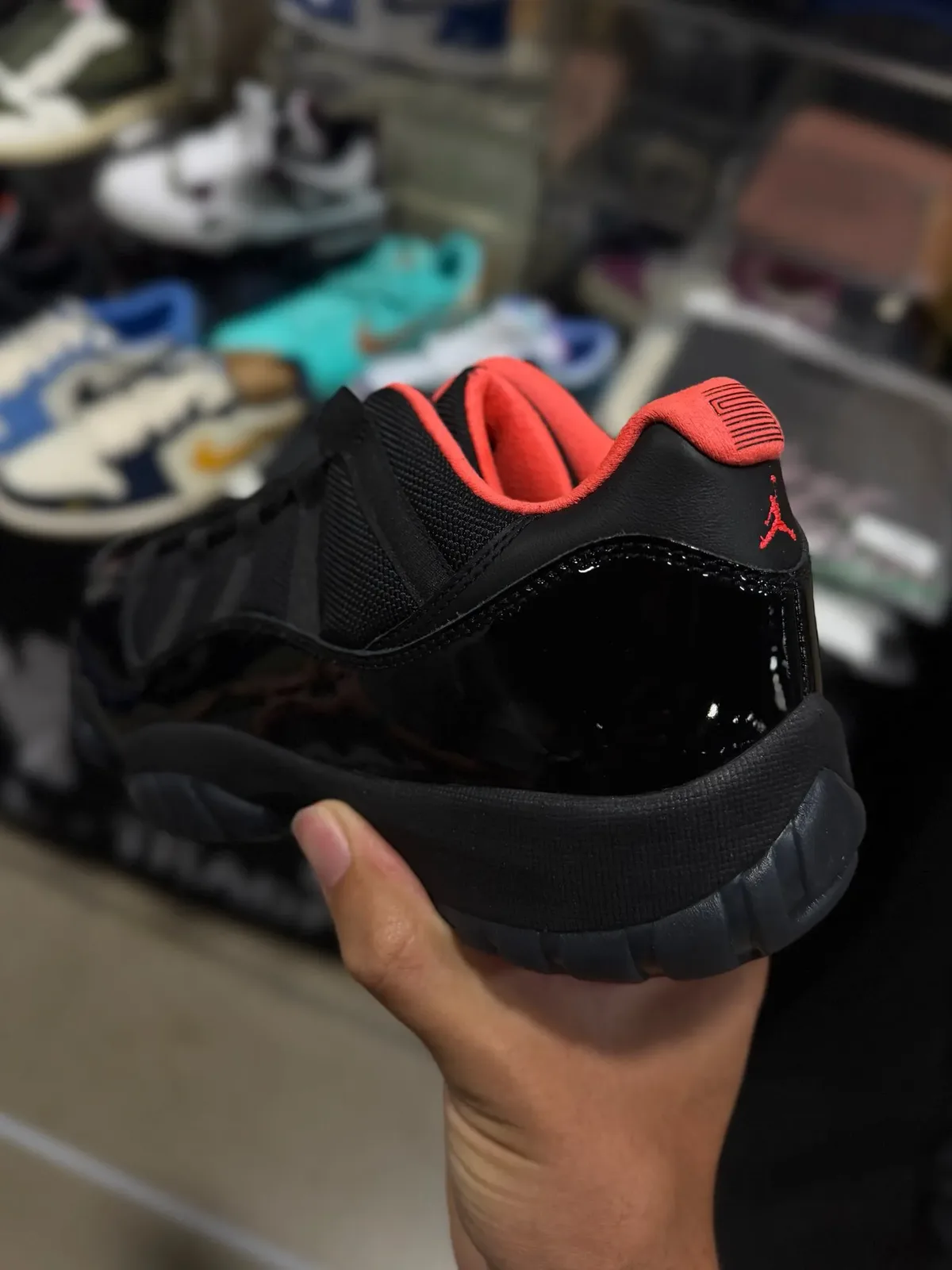 held in hand, heel angle with coral collar, red Jumpman logo, blurred sneaker shelves behind, Black Infrared