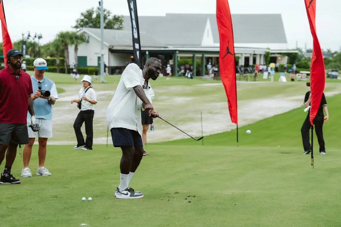 man swinging golf club near clubhouse with red Jumpman flags and onlookers, investment, no product)