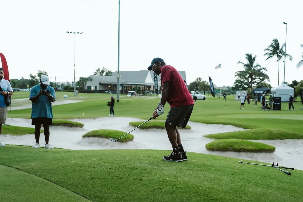 man mid-swing beside sand bunkers with red flag, clubhouse and palm trees behind, investment, no product)