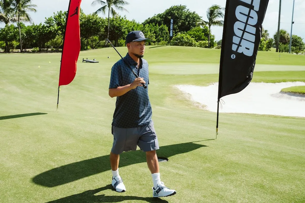 man walking on green holding golf club, red Jumpman flag and black 'tour' flag behind, investment, no product)