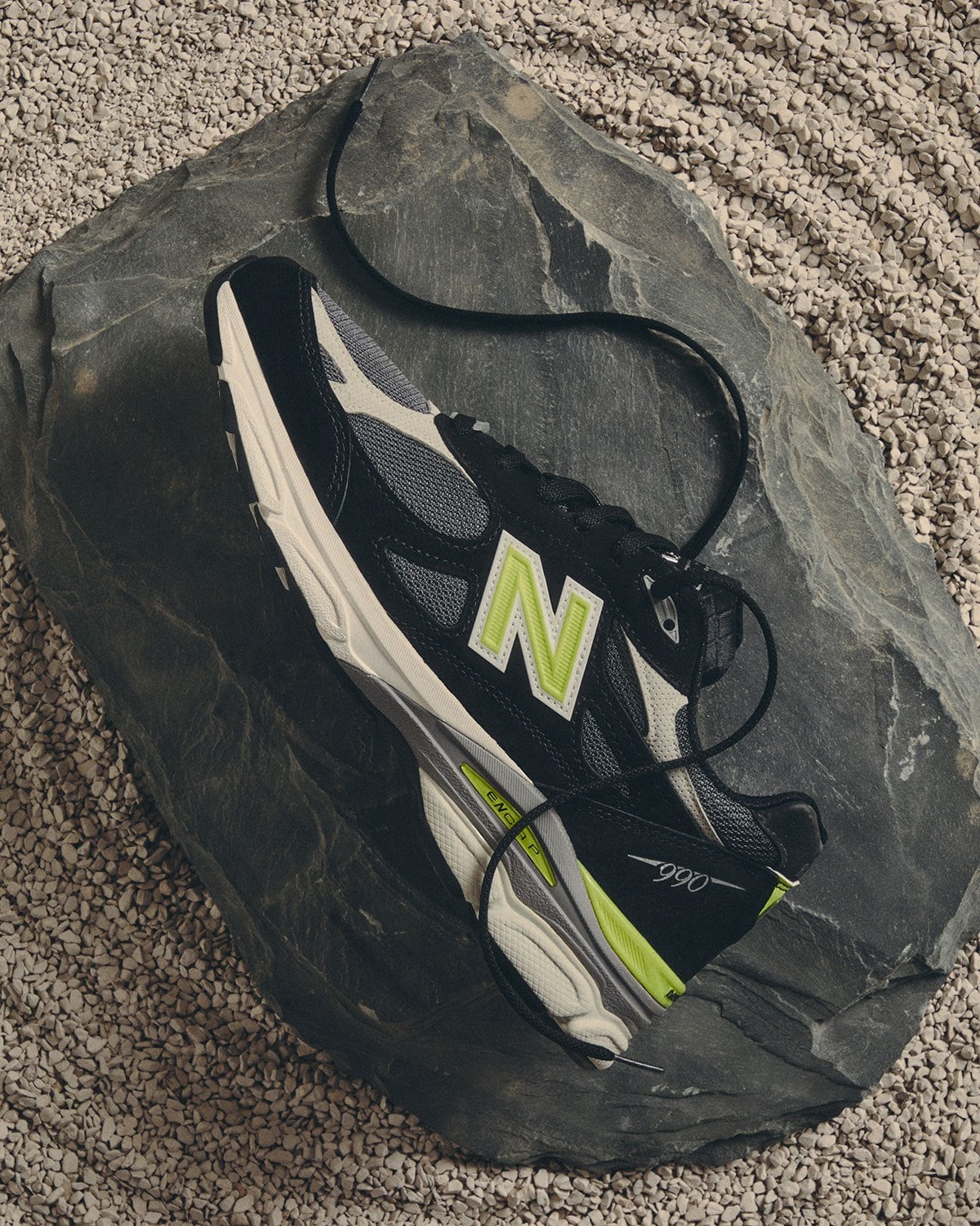 Top-down lateral view, black pair with lime accents, laces coiled on dark rock, New Balance 990v3