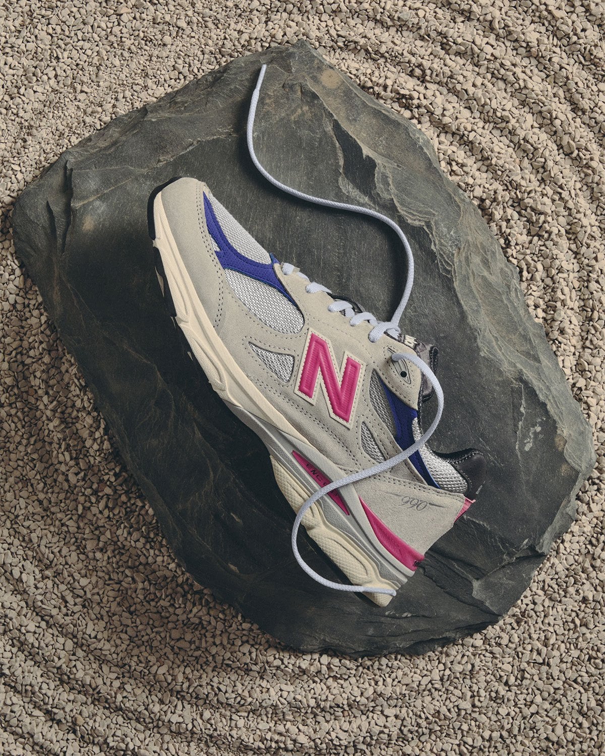 Top-down lateral view, grey/blue/pink pair with laces coiled on dark rock, New Balance 990v3
