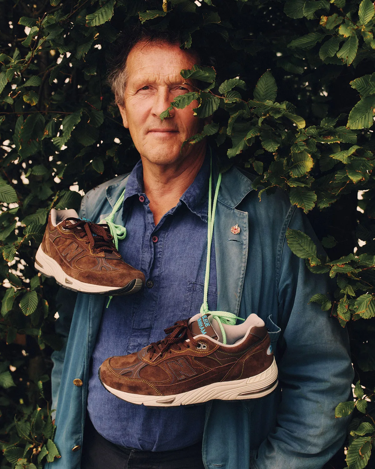 man holds pair by mint laces slung around neck, brown suede sneakers, x Bleue Burnham