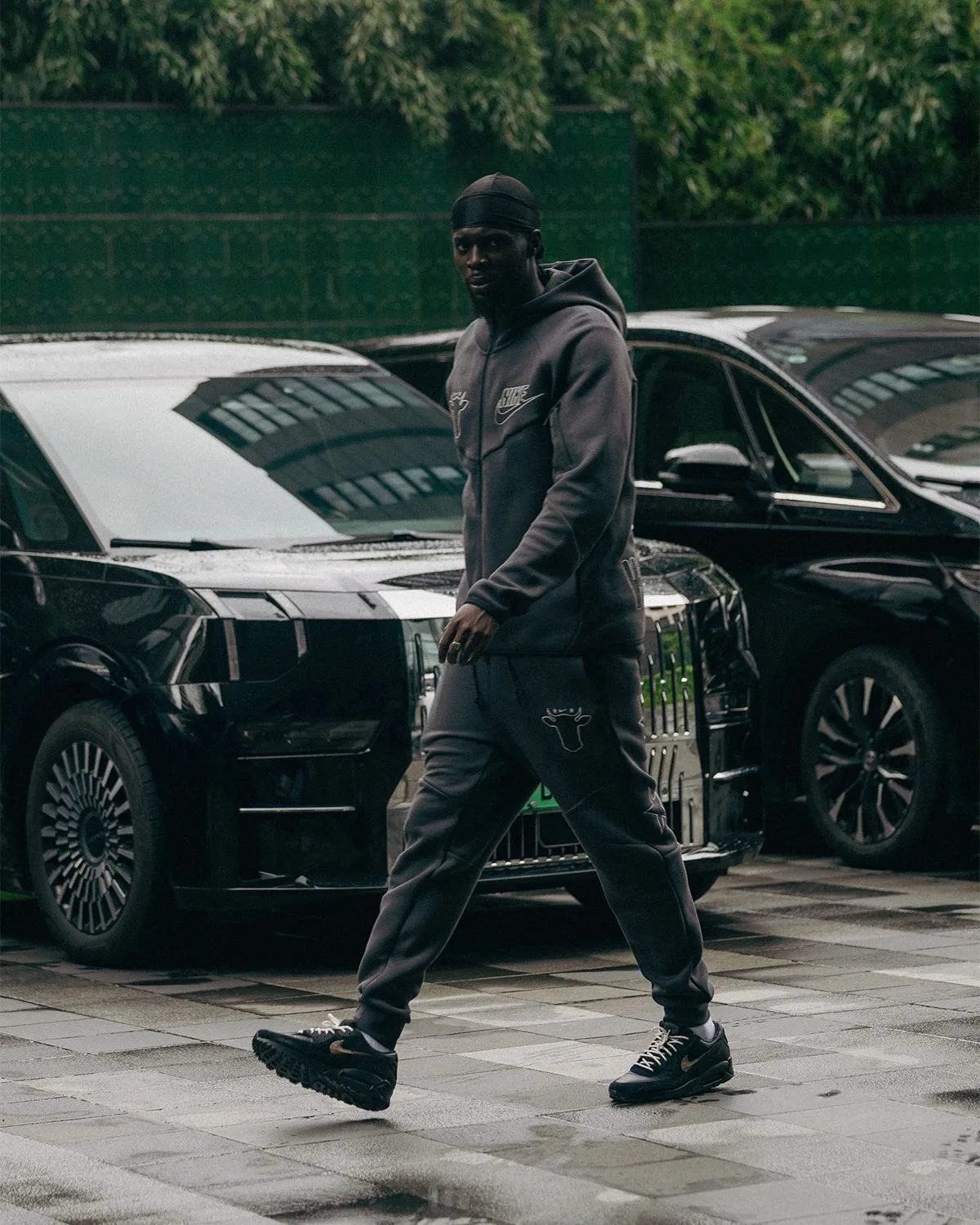 on-foot street view, dark tracksuit with cow-head logo and black pair beside parked cars, 90 x Wagyu
