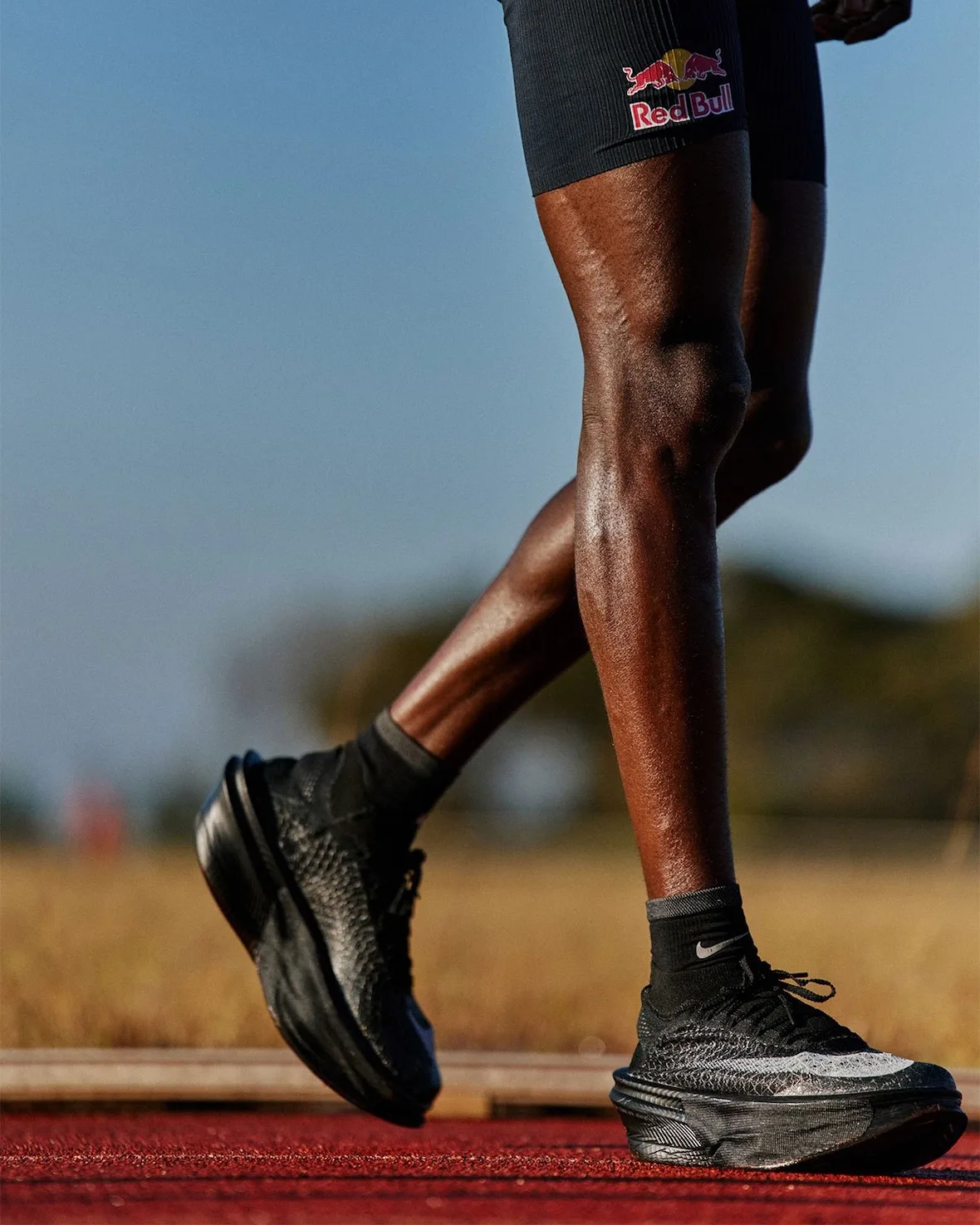 on-foot side view of a runner's legs mid-stride in black shoes, red bull shorts, Nike Apex