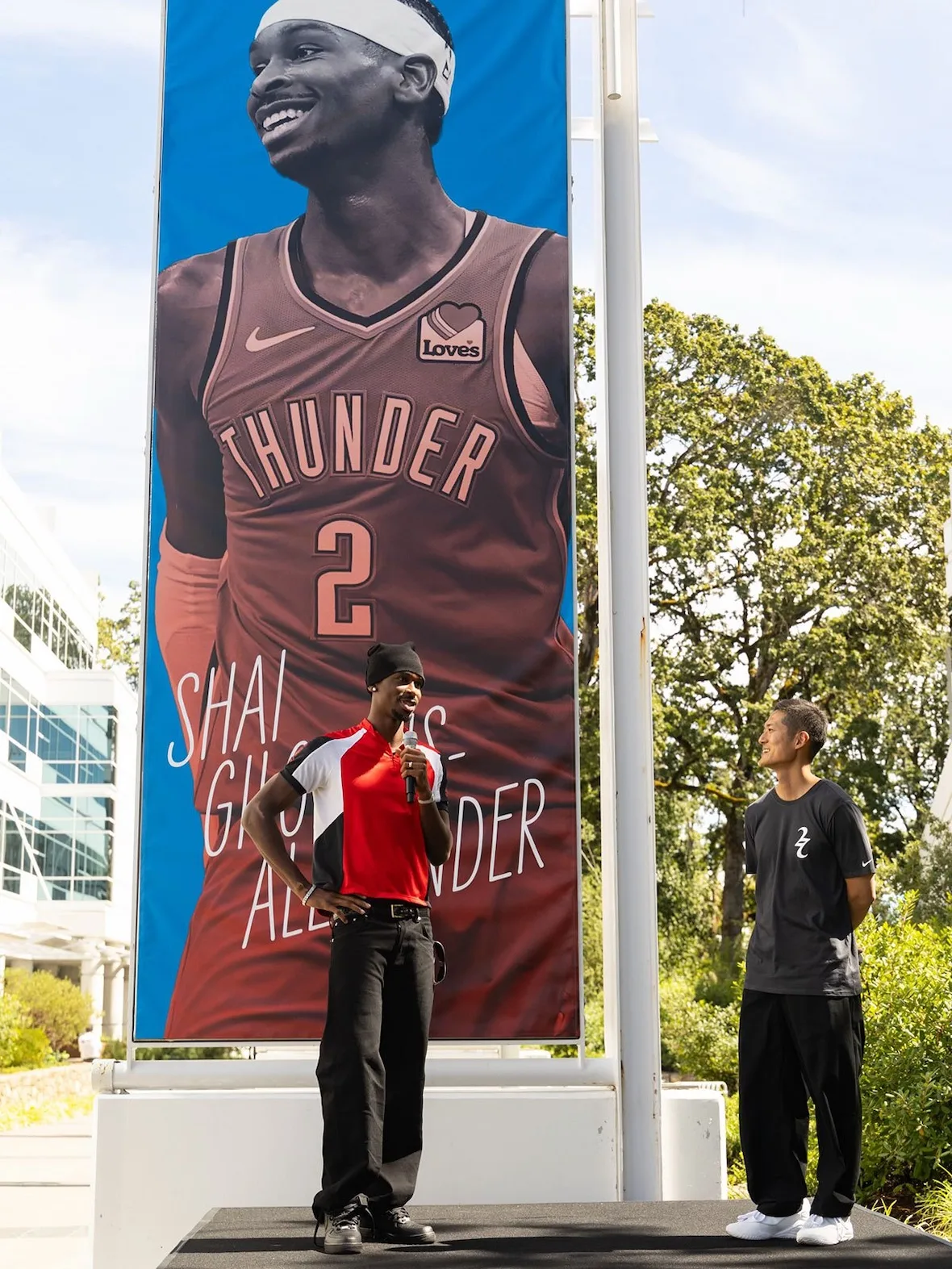 Two men on a stage, orange jersey and black shirt, large player banner behind, Nike SHAI 002
