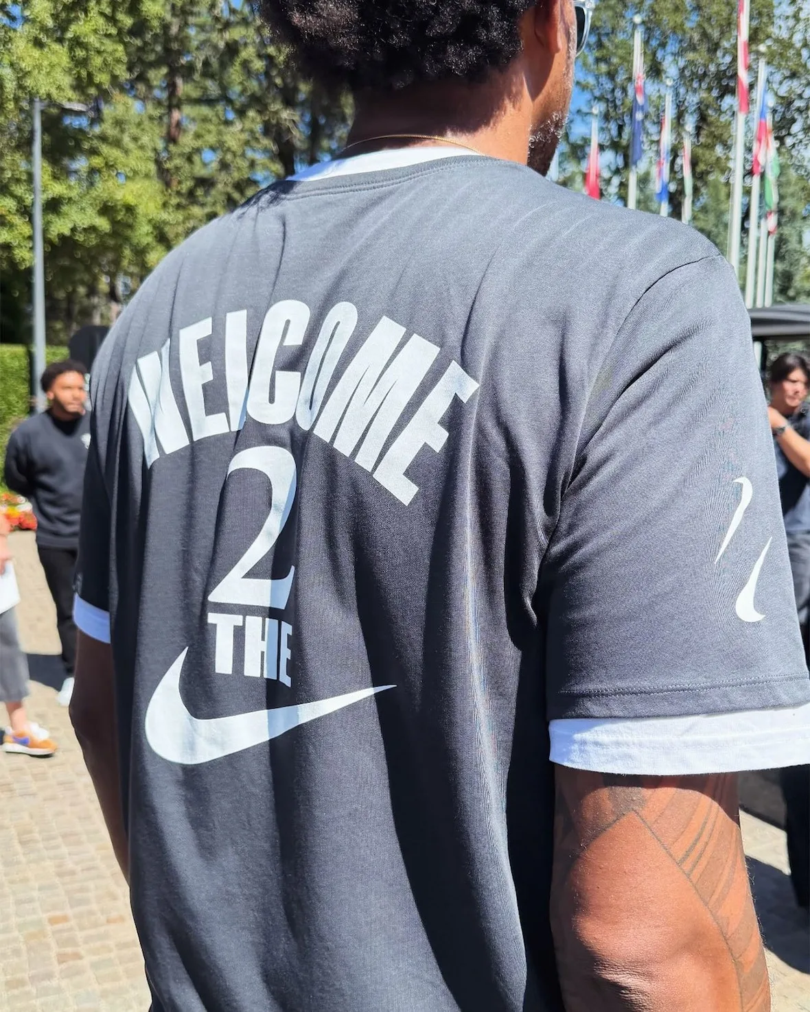 Close-up back of a grey t-shirt reading 'Welcome 2 The' with Nike swoosh, tattoo, Nike SHAI 002