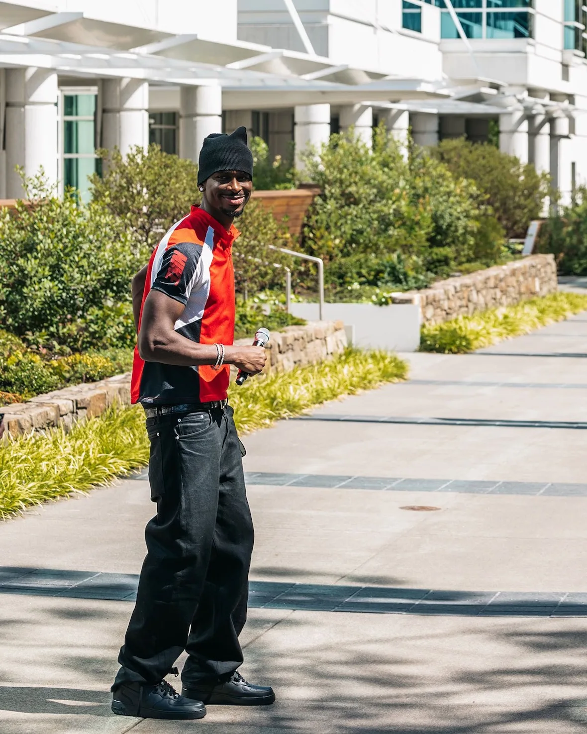 Full-body shot in an orange, white and black jersey, black jeans and sneakers, courtyard, Nike SHAI 002