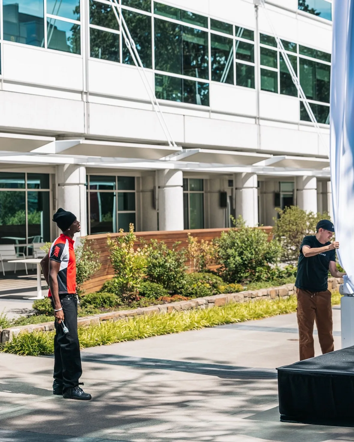 Two men in a courtyard, one in an orange and black jersey, glass office building behind, Nike SHAI 002