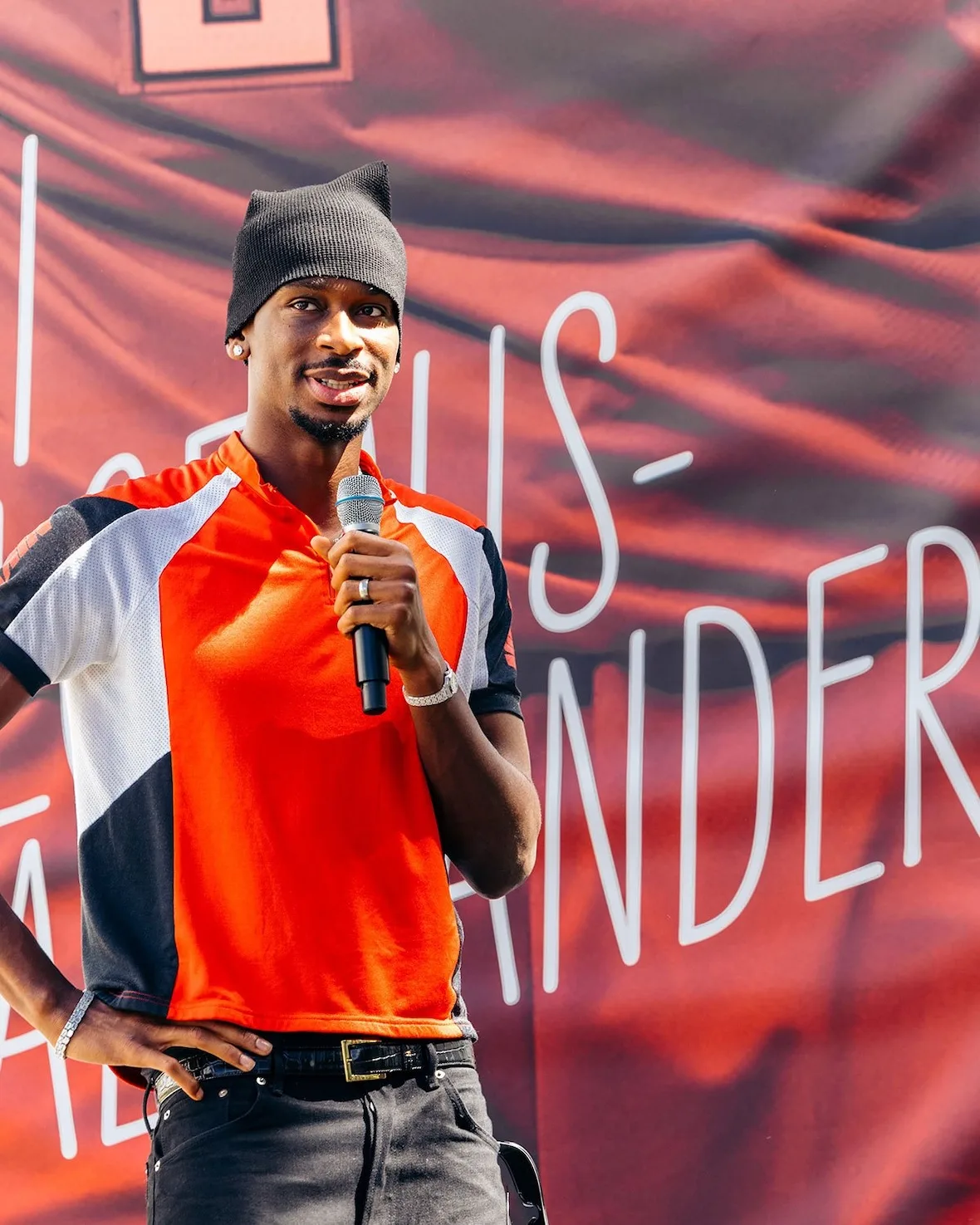 Close-up torso holding a microphone, orange jersey, red 'Alexander' banner behind, Nike SHAI 002