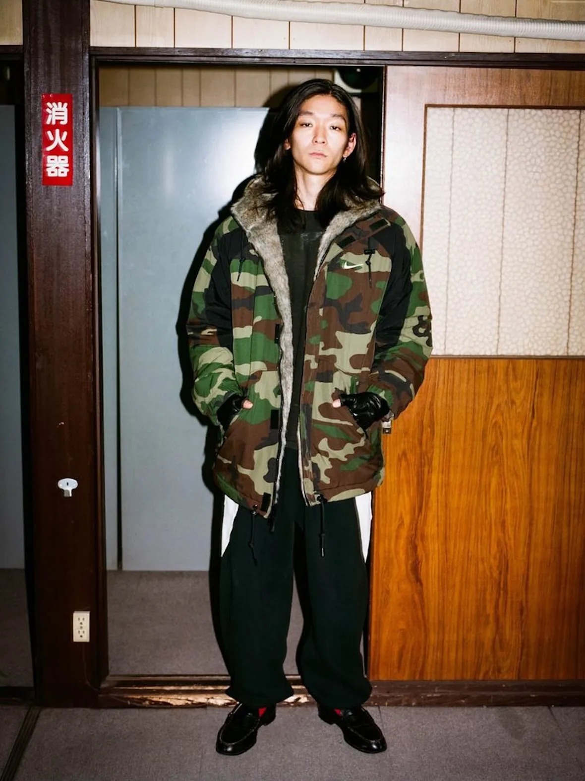 front view, camo parka with fur-lined hood, black gloves, wood-paneled hallway, Fall 2026 Collection