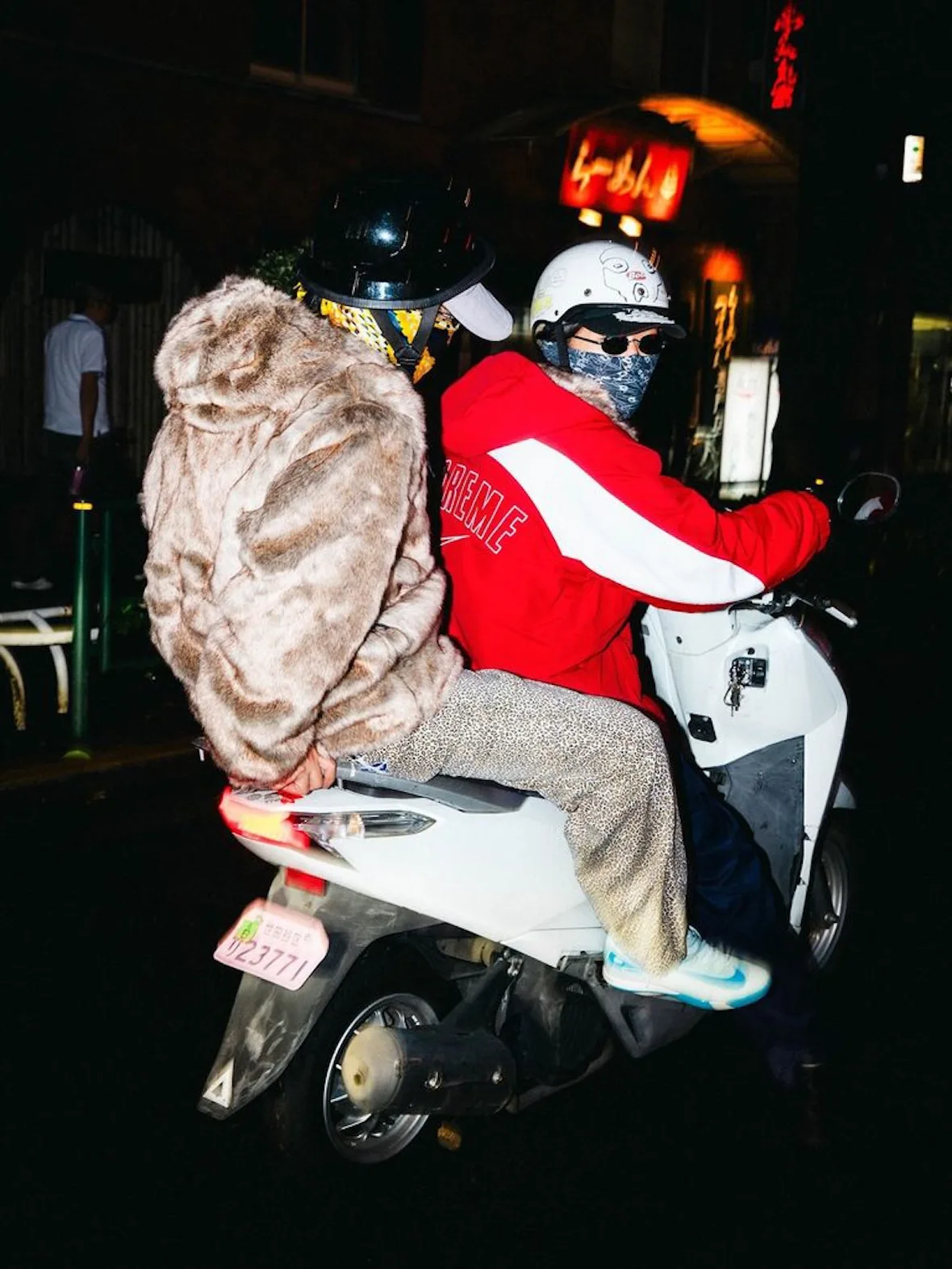 on-foot pair on a scooter, red Supreme jacket and tan faux-fur hooded jacket, night, Fall 2026 Collection