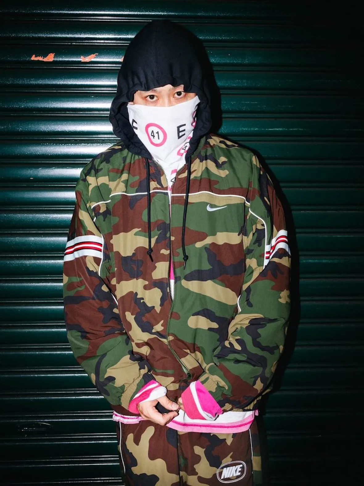 front view, camo hooded jacket with Nike swoosh, red-striped sleeves, bandana mask, Fall 2026 Collection