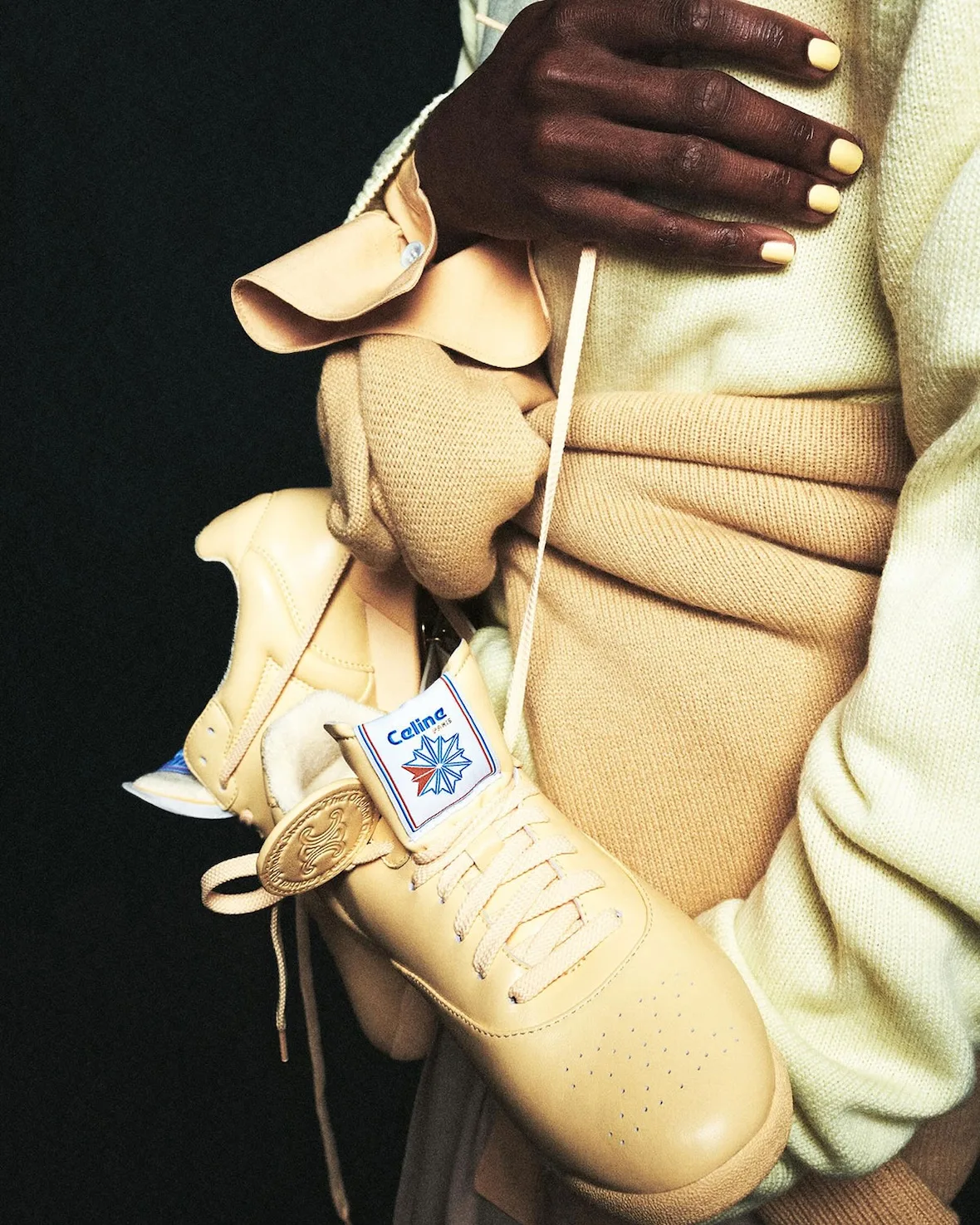 In-hand close-up, tan leather sneaker held against torso, Lo x Celine