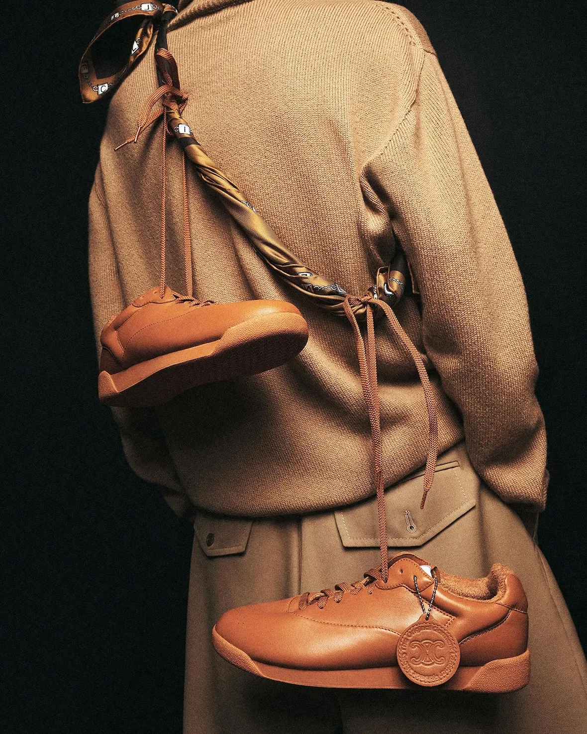 In-hand pair, tan leather sneakers dangling from a silk scarf strap, Lo x Celine