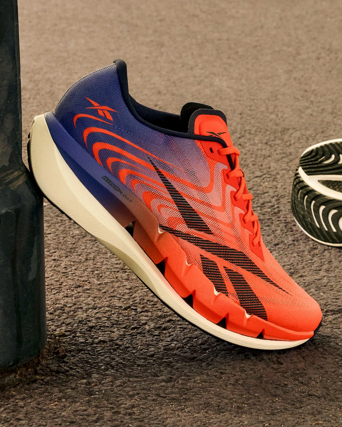 angled lateral view resting against a pole outdoors, orange and blue upper on asphalt, Reebok SpeedShifter