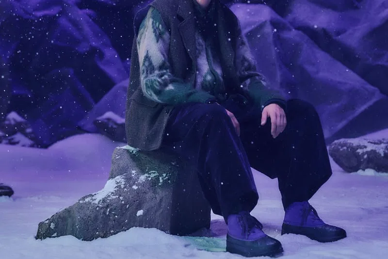 Seated model on rock detail shot showing purple duck boots and dark trousers, NEEDLES Ambler Collection