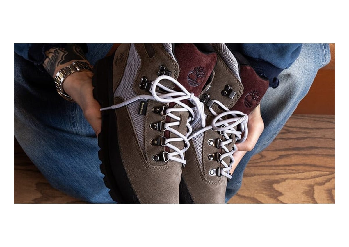Held overhead in hands, taupe suede boot with burgundy tongue and white laces, Timberland By Victor Kan F26 Euro Hiker GTX