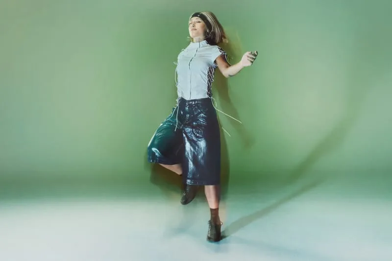 on-foot, model mid-jump in a denim skirt and dark boots, green backdrop, Tomorrowland WR1