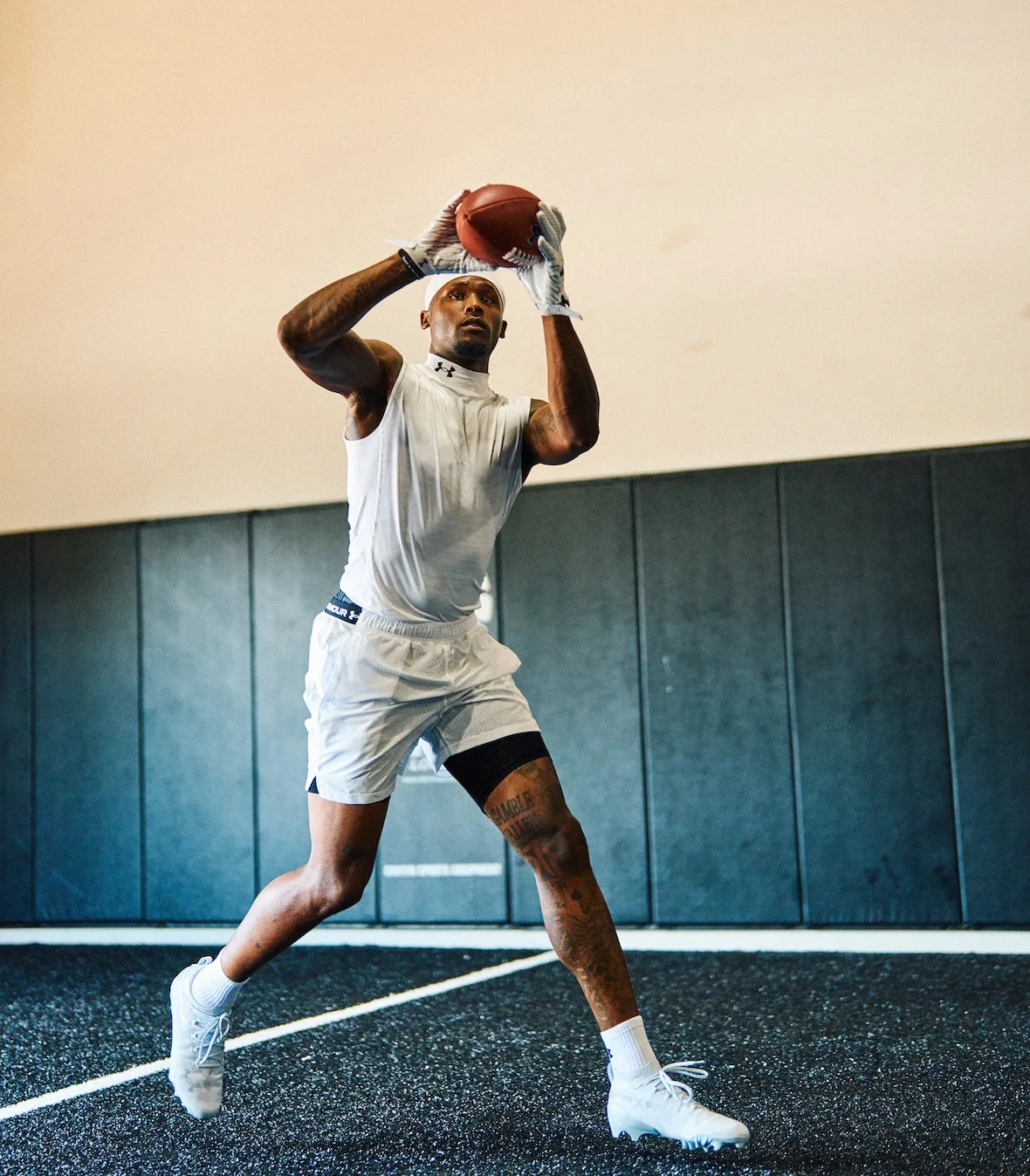 running catch of a football, white tee and shorts, cleats, black padded wall, x Under Armour