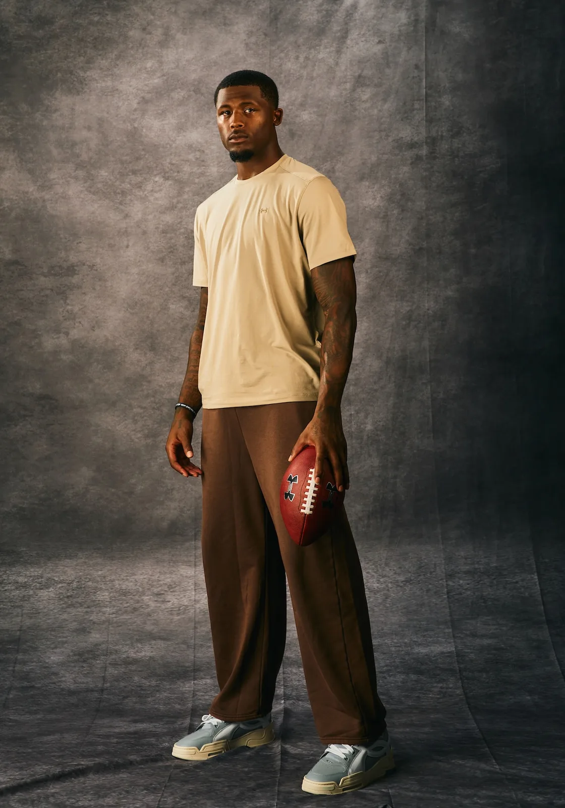 full-body studio shot, cream tee, brown pants, football, grey-cream sneakers, x Under Armour