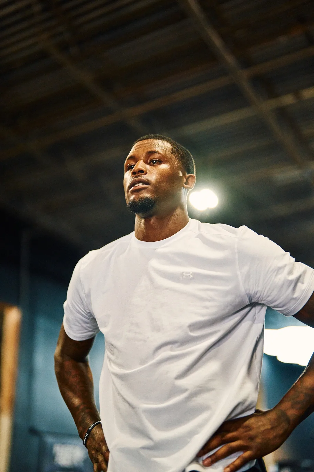 low-angle shot, white Under Armour tee, hands on hips, dim warehouse rafters, x Under Armour
