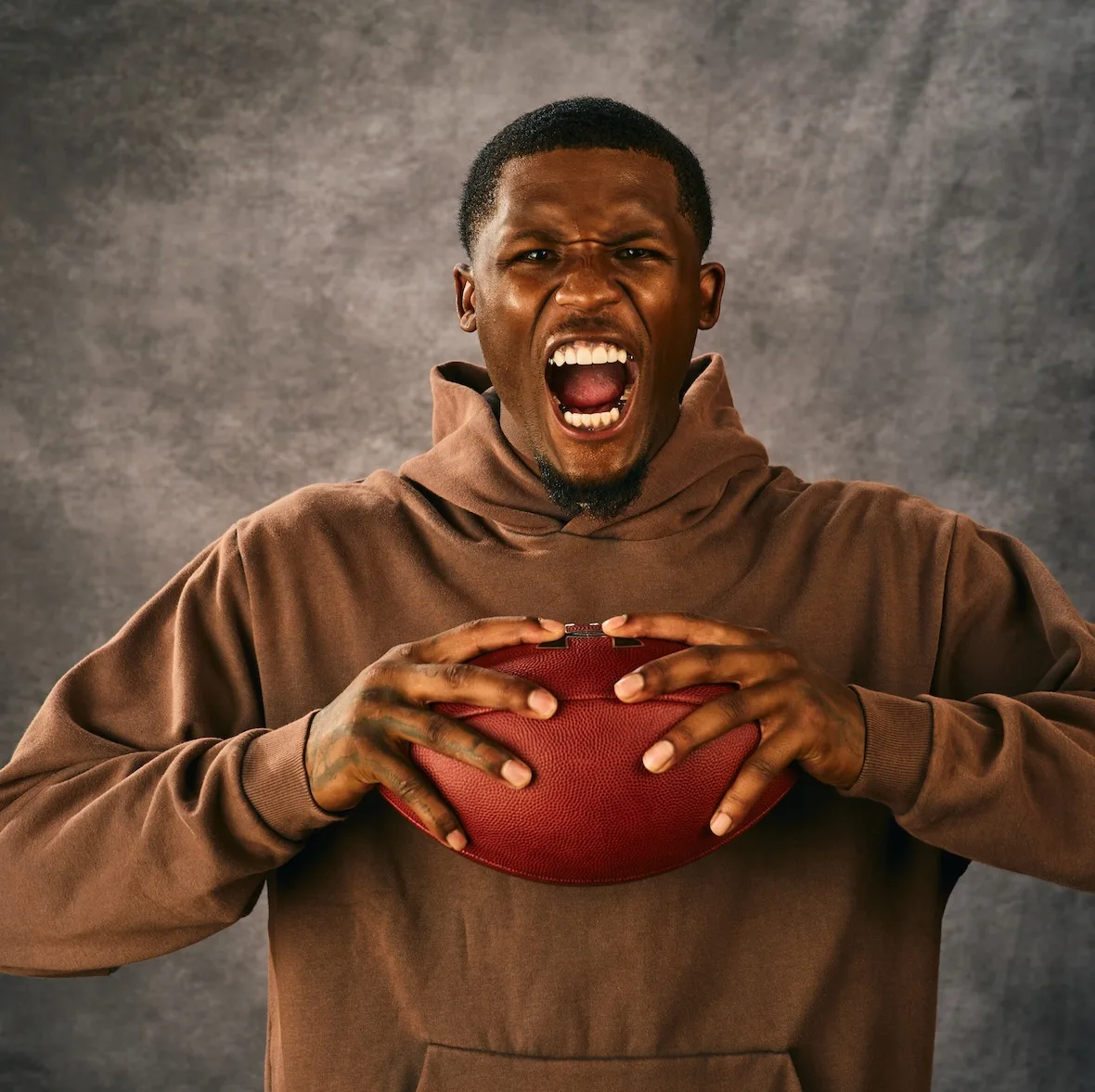 chest-up shot shouting, brown hoodie, gripping a football with tattooed hands, x Under Armour