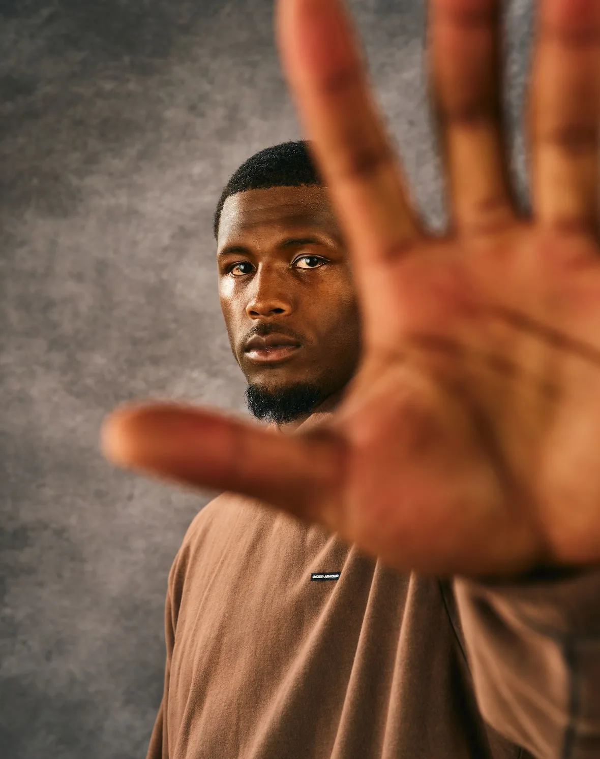 close-up face with blurred hand in foreground, brown tee with UA tag, x Under Armour