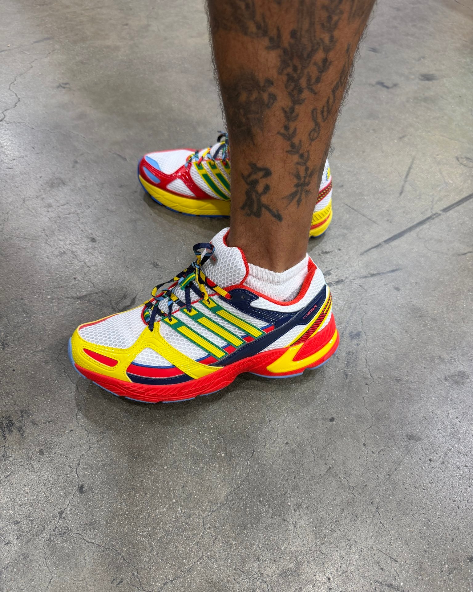 On-foot lateral view with tattooed leg, white mesh, yellow overlays and red sole on concrete, AdiStar Control 2