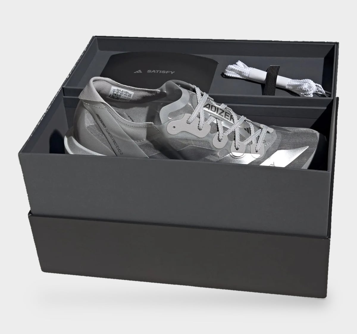 Shoe in open dark grey box with black SATISFY card and white spare laces, 2 x Satisfy