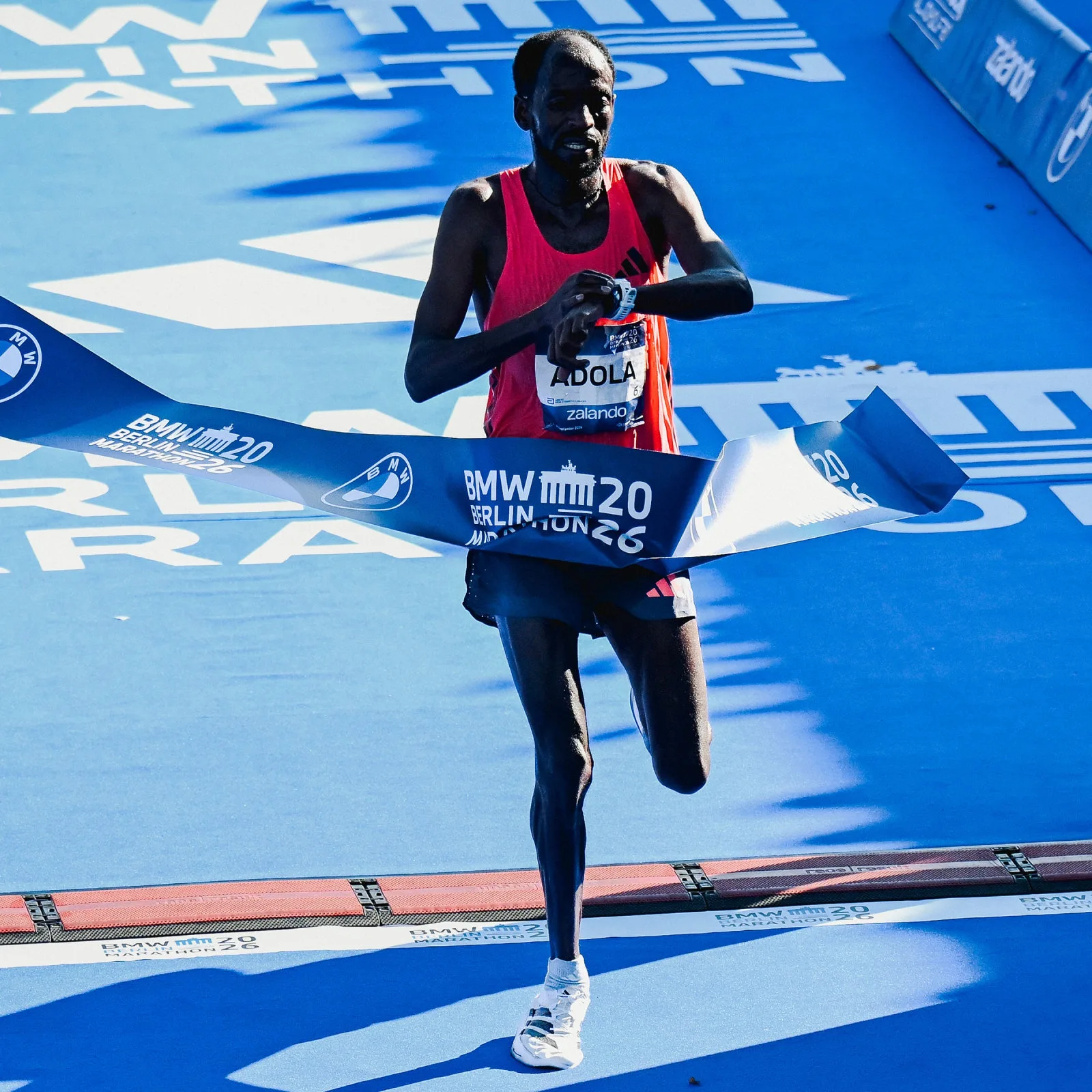 Runner in red vest crossing a BMW Berlin Marathon finish tape on blue ground, checking watch, Pro EVO 3