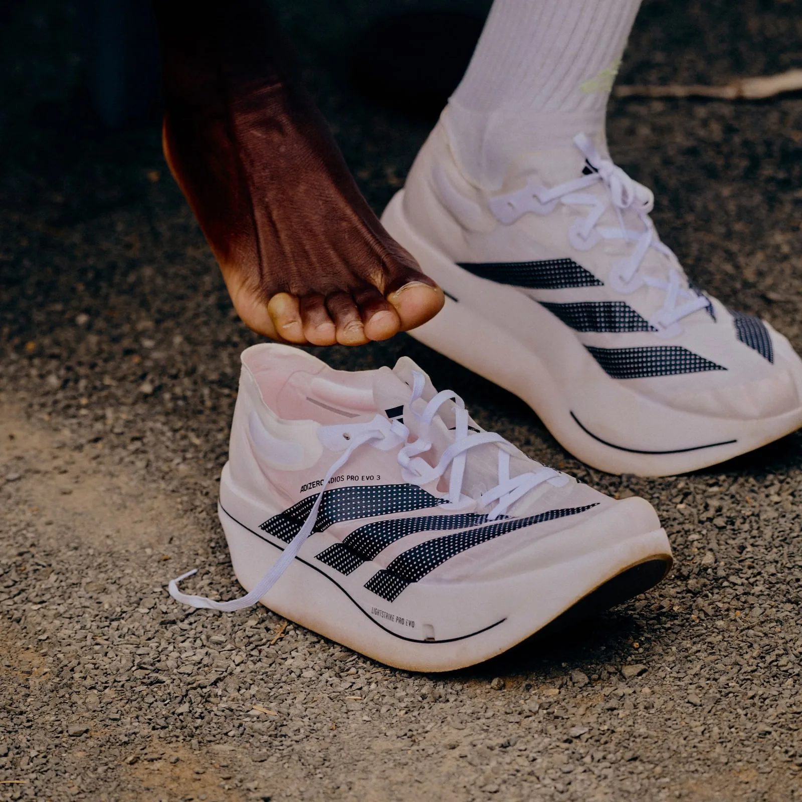 Barefoot foot over a white shoe with black dotted stripes and loose laces on gravel, Pro EVO 3