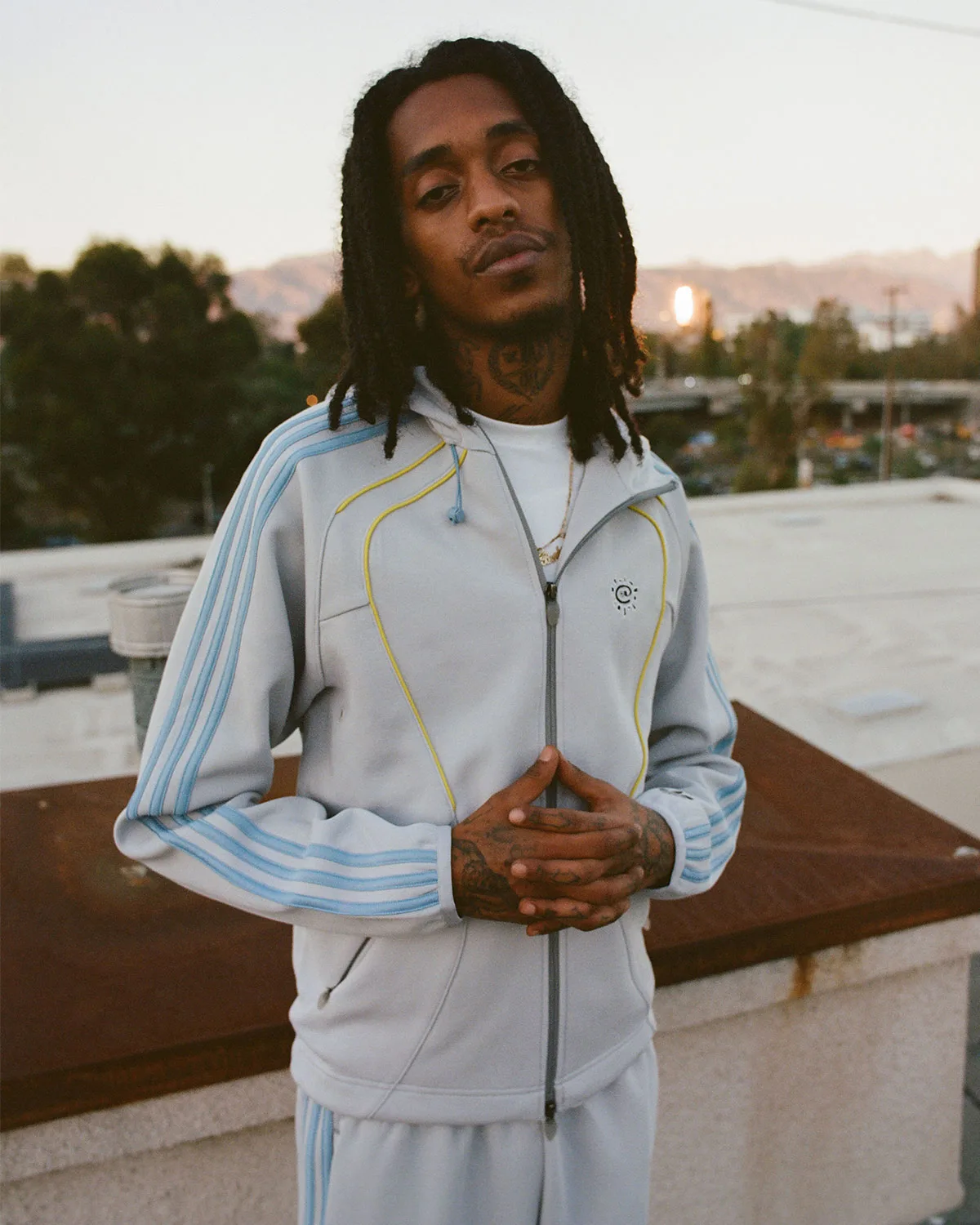 Person in pale grey track jacket with blue stripes and yellow piping, standing on a rooftop, Climacool x ADWYSD
