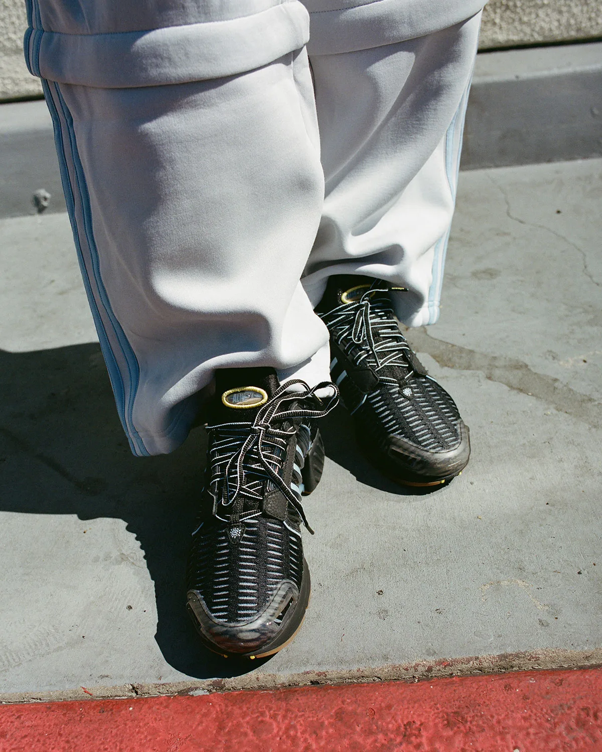On-foot view of black mesh sneakers with patterned laces, worn with pale grey striped pants on concrete, Climacool x ADWYSD