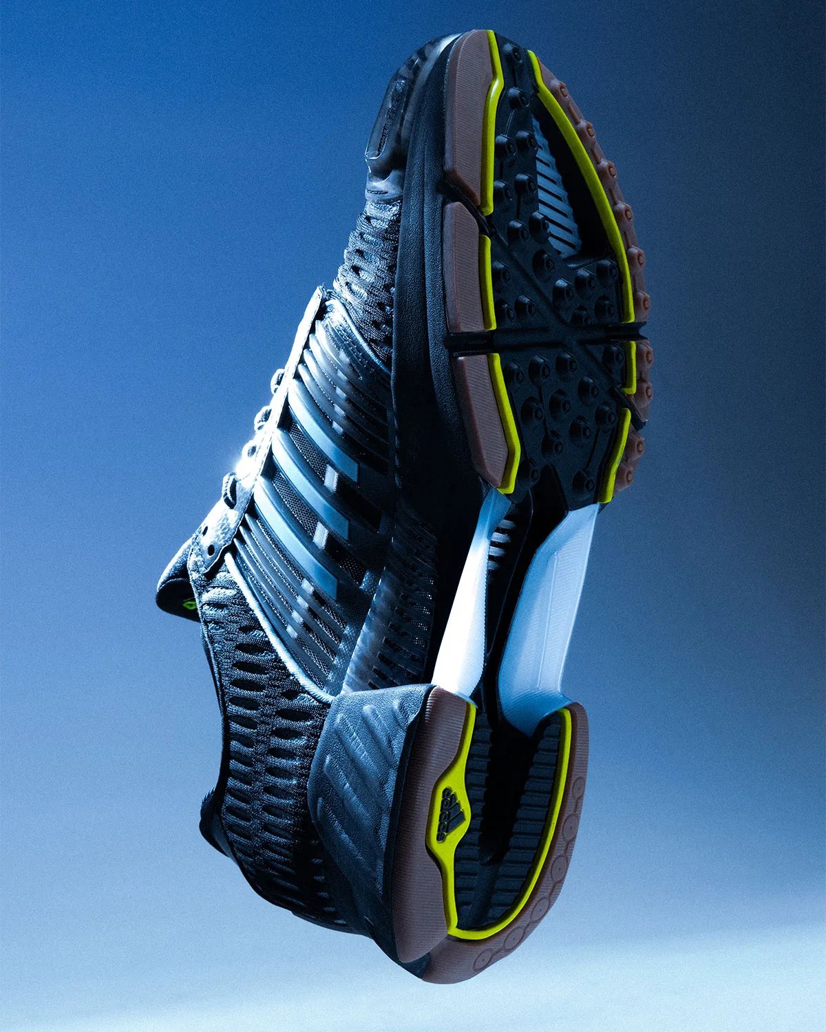 Outsole view of black sneaker with yellow-green trim, gum rubber and light blue midsole on blue, Climacool x ADWYSD