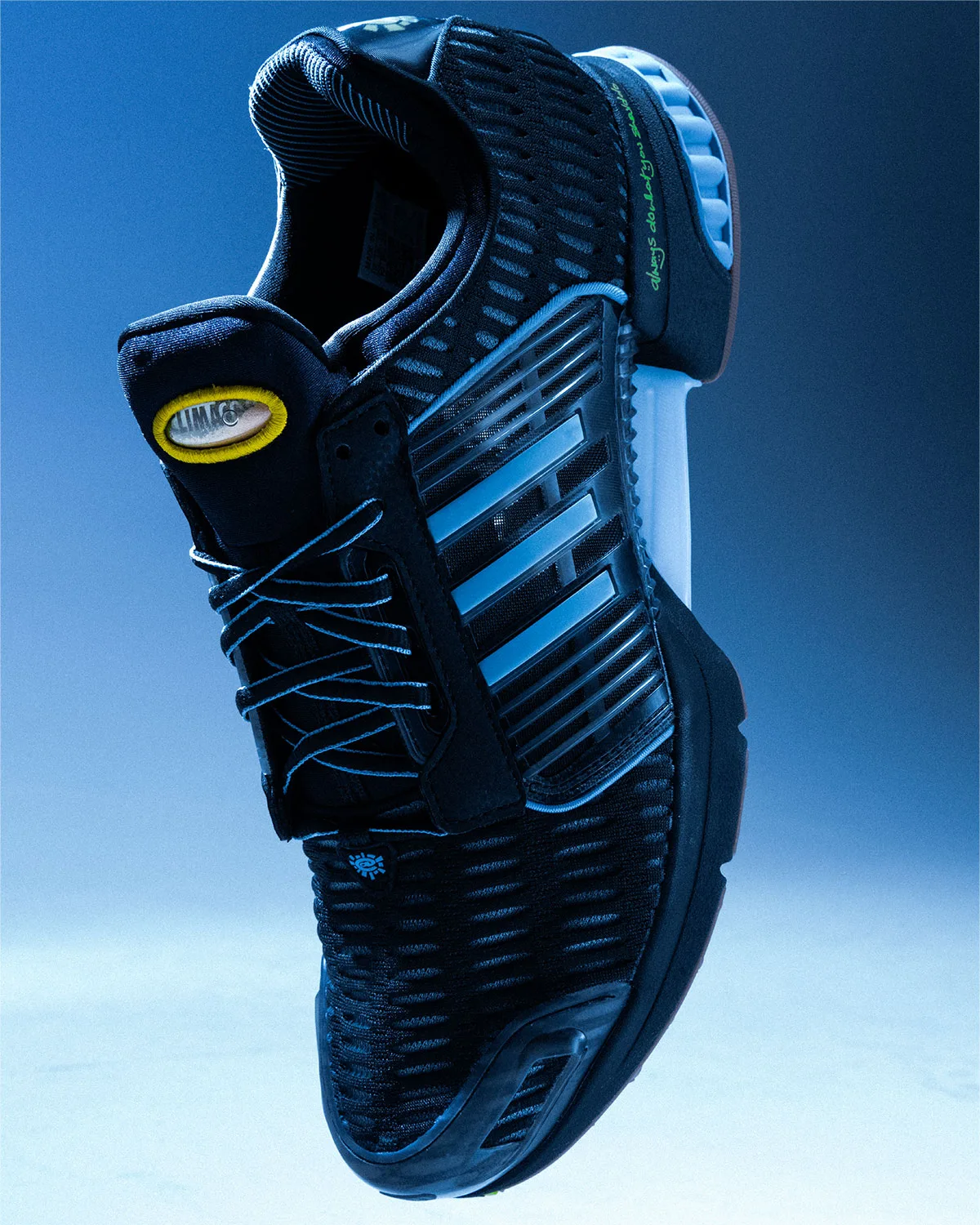 Top-down angled view of black mesh sneaker with blue stripes, yellow oval tongue tag, blue backdrop, Climacool x ADWYSD