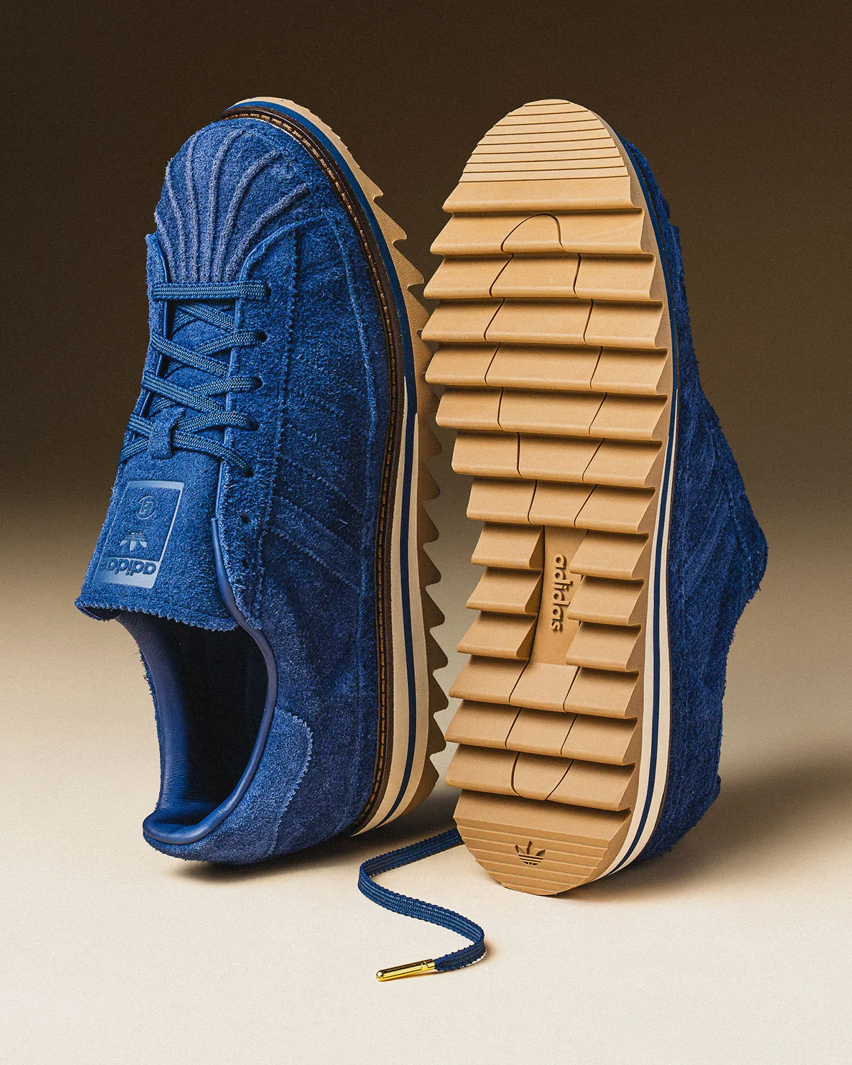 Blue suede sneaker beside its tan ridged outsole with adidas text, navy lace with gold tip, Mule x CLOT