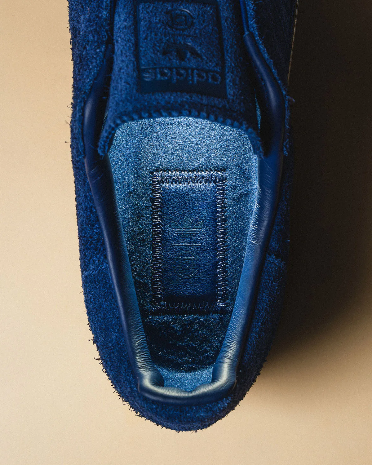 Top-down detail inside blue suede shoe showing leather-patched insole with trefoil and logo, Mule x CLOT