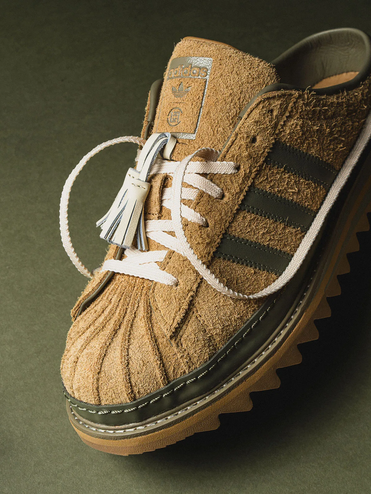 Angled close-up of tan suede toe, olive stripes, cream tassel laces and tongue label on olive, Mule x CLOT