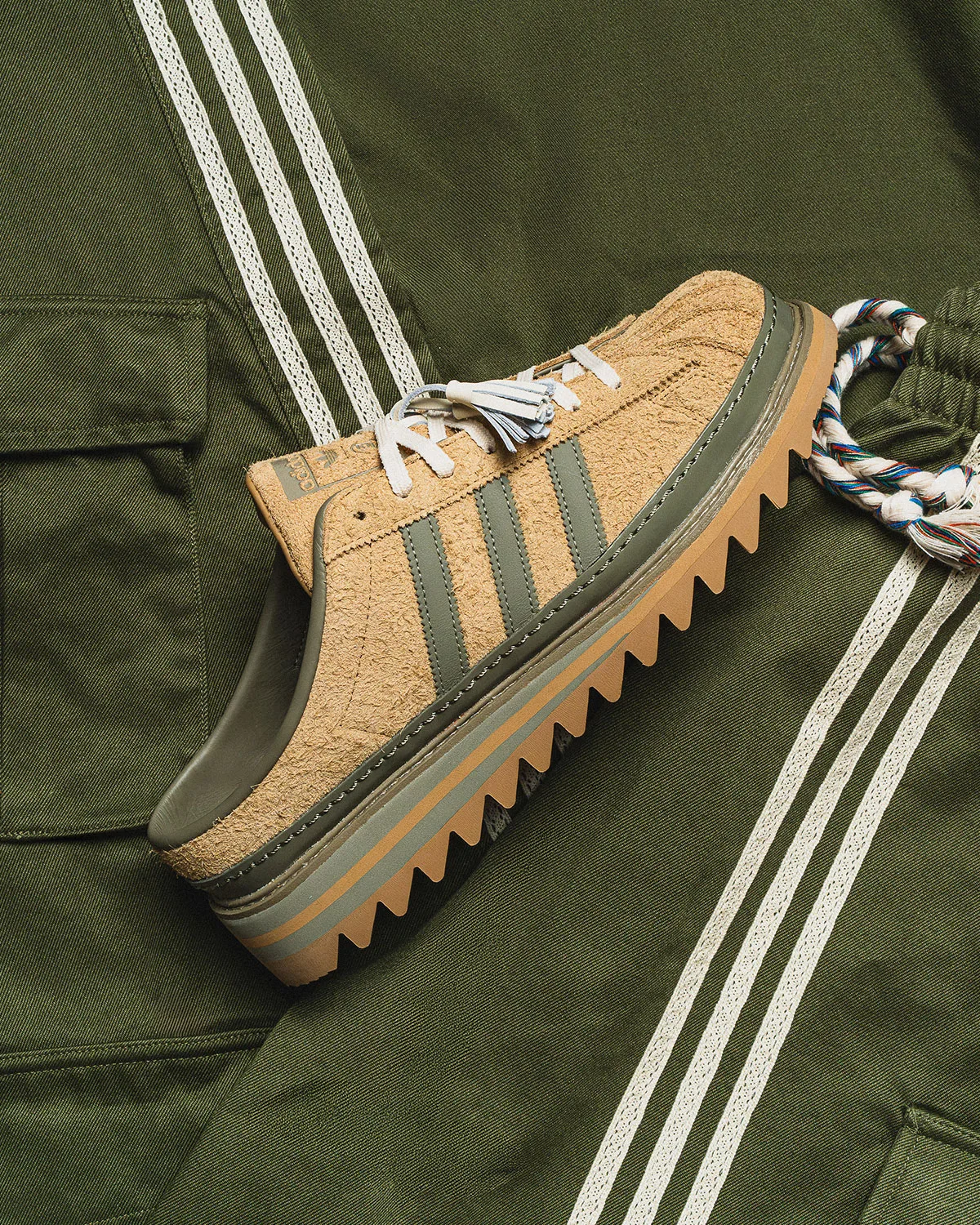 Lateral view of tan suede mule with olive stripes, tassel laces and sawtooth sole on olive fabric, Mule x CLOT