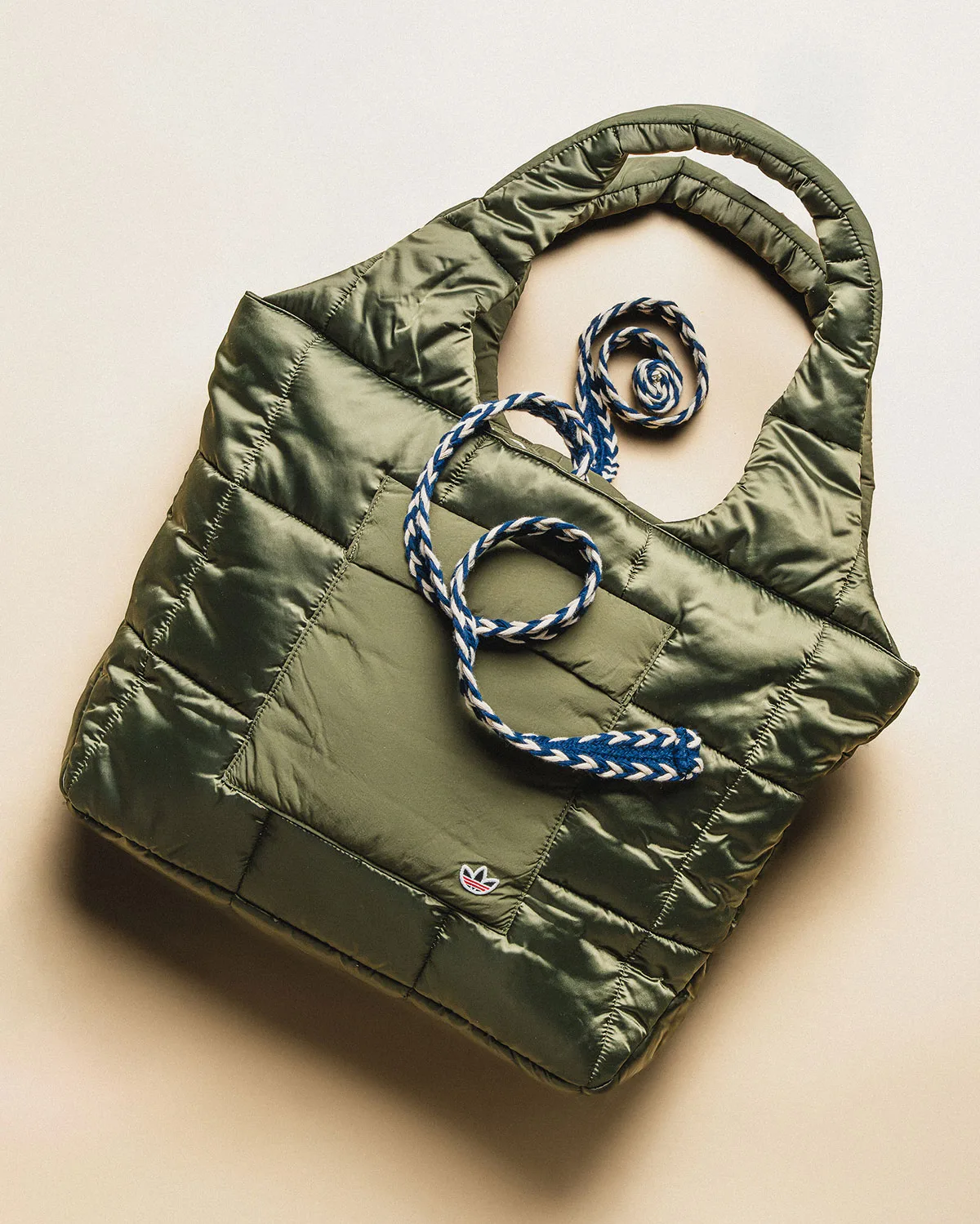 Padded olive nylon tote bag with blue and white braided cord and small trefoil patch, on beige, Mule x CLOT
