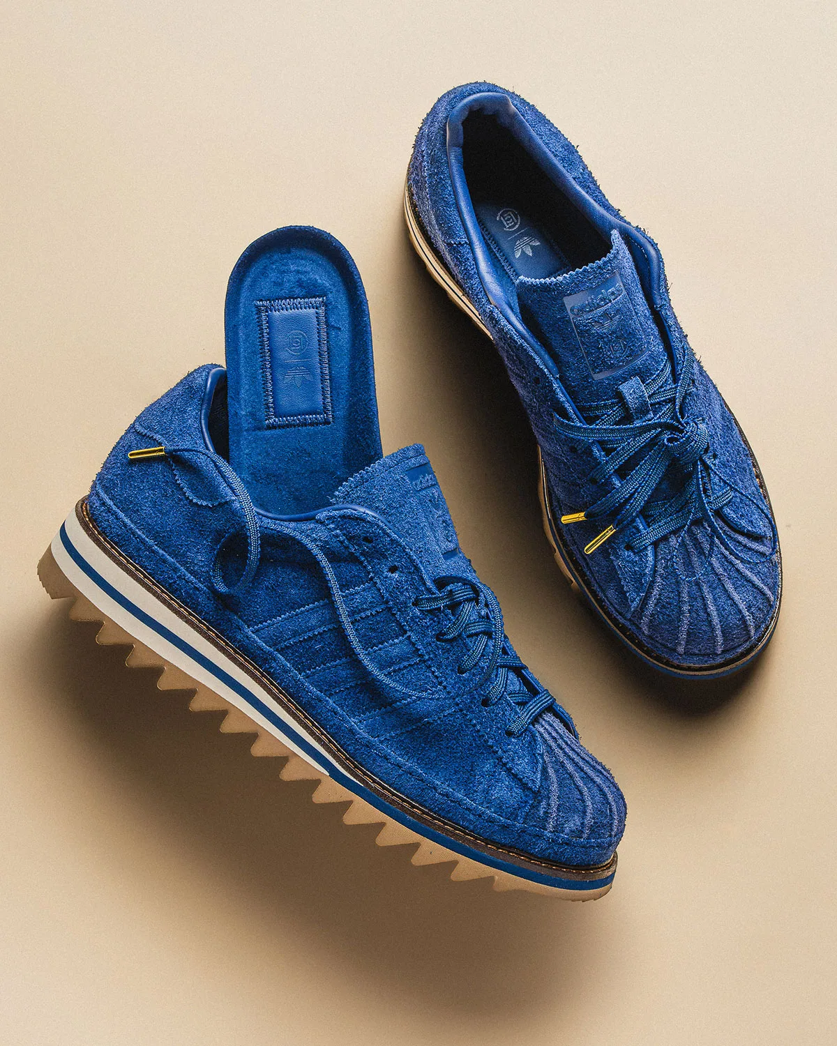 Top-down pair of blue suede sneakers with gold lace tips, one showing a blue insole, on beige, Mule x CLOT
