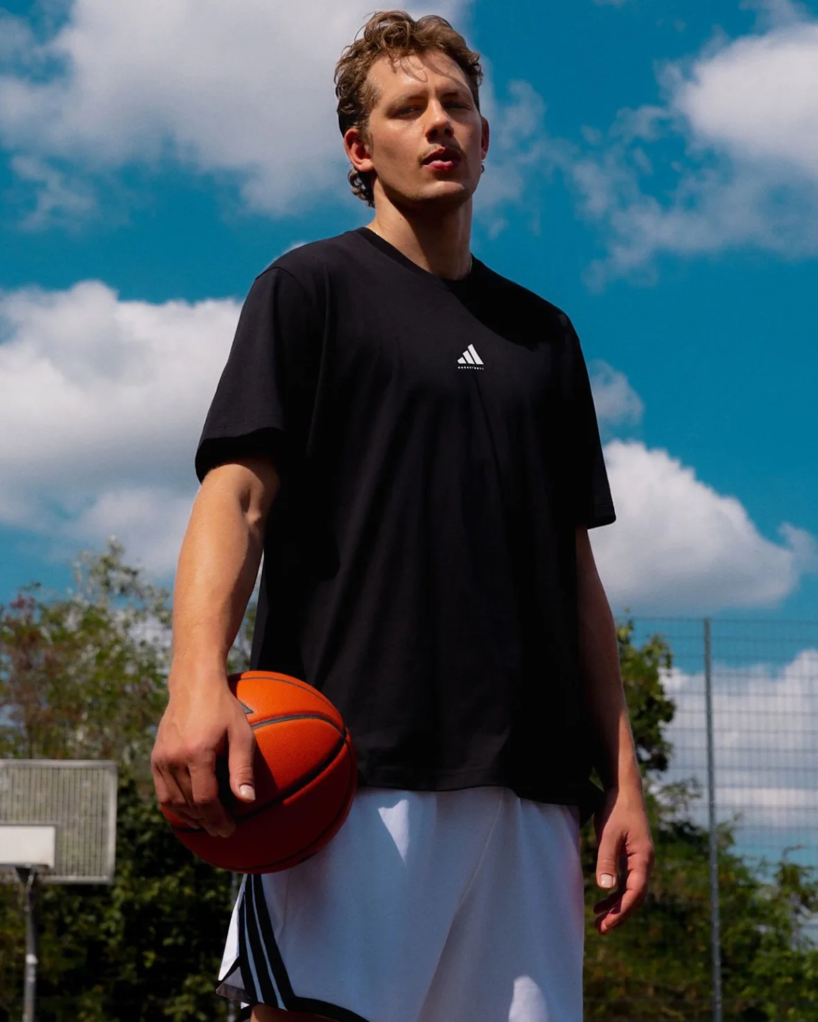 On-court portrait of a man in a black adidas tee holding a basketball under a blue sky, Wagner Brothers
